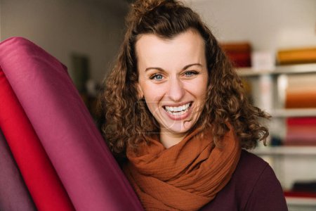 Joyous young woman with curly hair and a radiant smile, holding vibrant fabric, colorful shelves in the background
