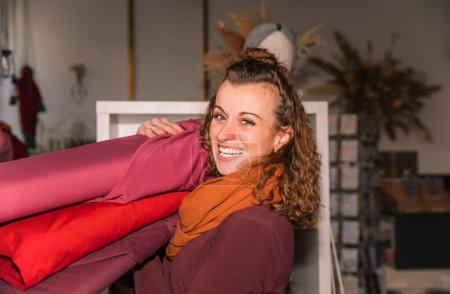 Friendly woman with curly hair in a creative workshop setting, handling a stack of vibrant fabric rolls