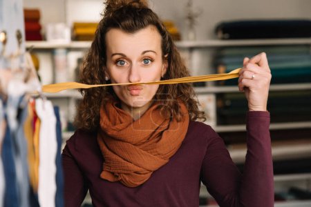 Playful moment captured of a curly-haired fashion designer balancing a material strip on her lip, showing her fun side in a shop