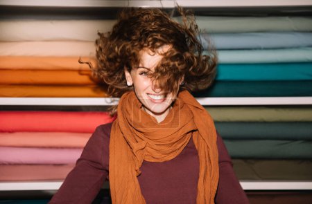 Playful young woman with curly brown hair smiling with her eyes closed in a vibrant textile shop