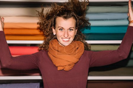 Mujer joven de pelo rizado sonriendo ampliamente, el pelo volando, en una tienda de tela de colores