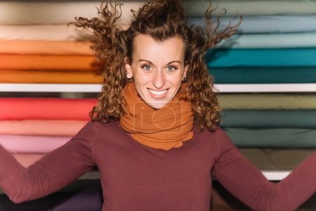 Une styliste exubérante célèbre joyeusement dans son atelier, les bras écartés, avec un fond de rouleaux de tissu colorés exprimant l'esprit vibrant de son espace créatif