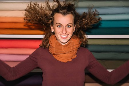 Una enérgica diseñadora de moda extiende sus brazos en alegre celebración, su cabello rizado bailando en el aire, contra un telón de fondo de coloridos rollos de tela en su estudio