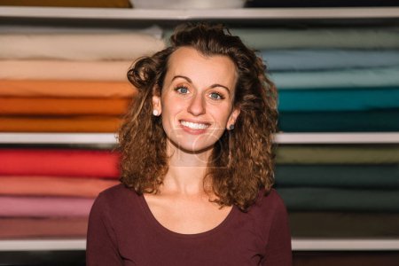 A confident fashion designer with curly hair poses in front of her well-organized colorful fabric rolls, exuding a professional and cheerful demeanor in her studio