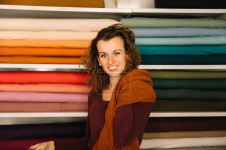 Gemütliches Porträt einer Modedesignerin mit lockigem Haar, das in einen weichen Schal gehüllt ist und ihr warmes Lächeln den Komfort und die Inspiration ihres mit Stoff gefüllten Ateliers widerspiegelt