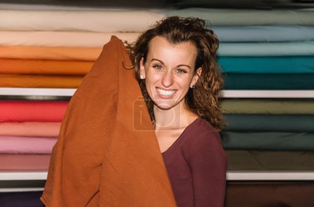Une créatrice de mode ludique avec un sourire éclatant s'enveloppe dans un grand morceau de tissu, son expression pleine de joie et de créativité dans le contexte coloré de son studio