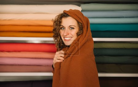 A smiling charming woman wrapped in a cozy scarf in a textile shop, design concept