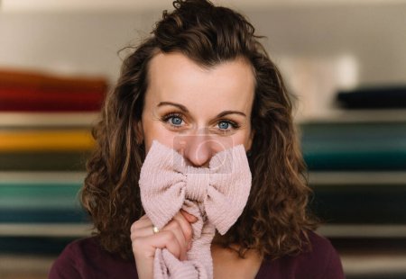 Close-up view capturing the vibrant eyes of a fashion designer playfully covering her face with a pink knitted bow, hinting at her creative personality in the backdrop of colorful fabric shelves
