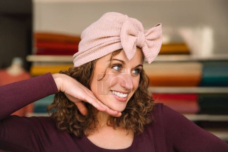 Joyful fashion designer playfully posing with a handmade pink bow headband, her hand as a pretend support, in her vibrant fabric-lined studio