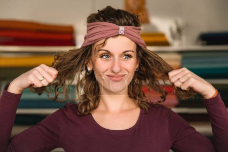Kreative Designerin trägt ein trendiges Haarband, während sie ihre lockigen Haare spielerisch anpasst, was durch die farbenfrohe Stoffkulisse ihrer lebendigen Werkstatt unterstrichen wird