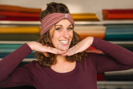 Eine verspielte Modedesignerin präsentiert ihre fröhliche Persönlichkeit und rahmt ihr Gesicht mit den Händen, während sie in ihrem lebendigen Textilatelier ein trendiges Haarband trägt