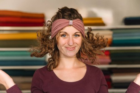Eine fröhliche Modedesignerin mit ausdrucksstarken Augen und lockigem Haar posiert verspielt in ihrem farbenfrohen Textilatelier und zeigt stolz ein schickes Haarband