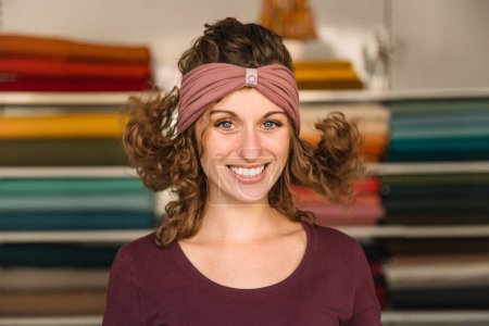Eine verspielte Modedesignerin mit lockigem Haar und funkelnden Augen präsentiert in ihrer Werkstatt ein lila Stirnband vor bunten Stoffrollen