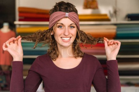 Verspielte Modedesignerin modelliert in ihrem Atelier ein mauvefarbenes Stirnband