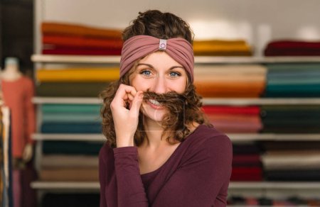 A cheerful fashion designer makes a playful mustache with her curly hair, wearing a stylish mauve headband, against a backdrop of colorful fabric rolls