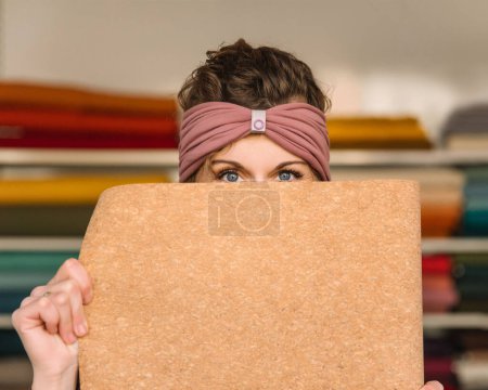 Curly-haired designer peeks playfully over a cork board, her eyes shining with creativity in her fabric-filled studio