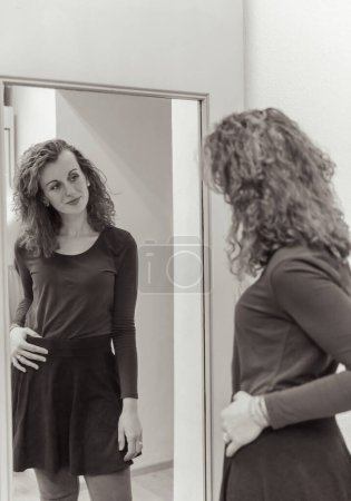 Stylish woman with curly hair looking at her reflection in a mirror, wearing a sleek black dress
