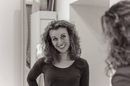 Lächelnde Frau mit lockigem Haar betrachtet ihr Spiegelbild in einem Ankleideraum-Spiegel