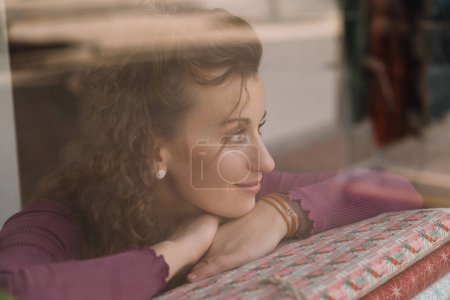 Woman with curly hair resting her chin on her hands, looking outside through a cafe window, captured in a reflective mood