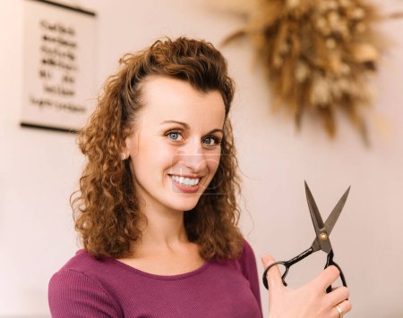 Cheerful young woman with curly hair holding a pair of scissors, ready to start a new project in her creative workshop