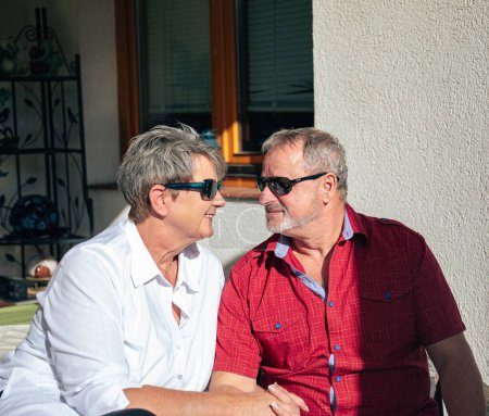 Téléchargez les photos : Femme souriante et homme en lunettes de soleil se regardant avec tendresse sur leur véranda - en image libre de droit