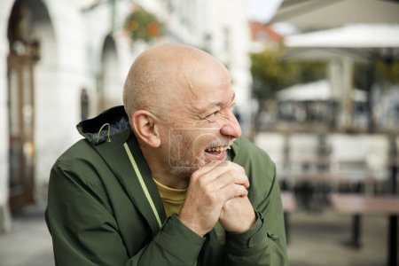Elderly bald man with beard, laughing in a green Marmot jacket, seated at an urban cafe during the day