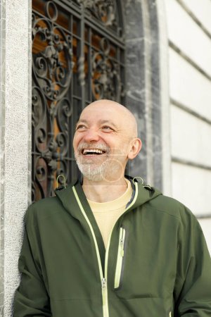 Zufriedener älterer Mann mit Glatze und Bart, der eine grüne Murmeltierjacke trägt und in einer sonnenbeschienenen städtischen Umgebung lächelt