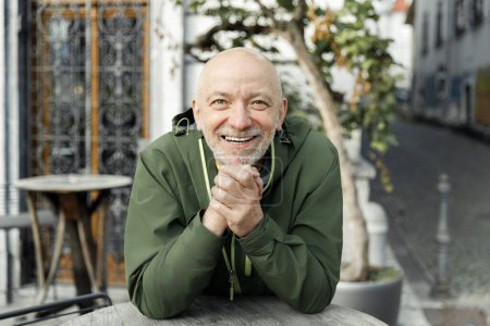 Happy senior man with a beard, clasping his hands joyfully while sitting outdoors in a green Marmot jacket