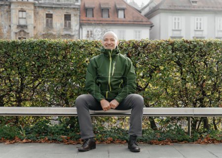 Smiling senior man sitting comfortably on a park bench in a green Marmot jacket and gray pants, surrounded by autumn foliage