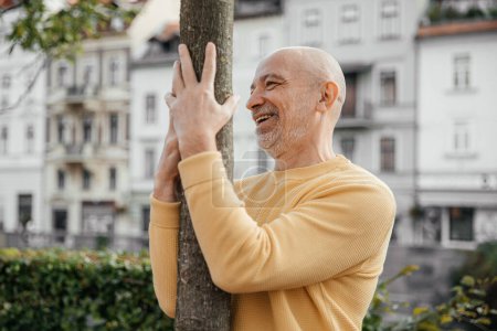 Senior Man in Yellow Sweater Laughing While Holding onto a Tree in Urban Park