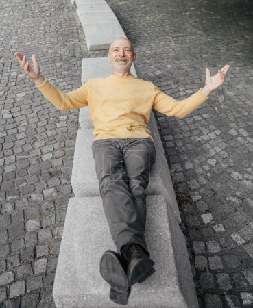 Älterer Mann im gelben Pullover lehnt mit weit geöffneten Armen auf einer urbanen Bank
