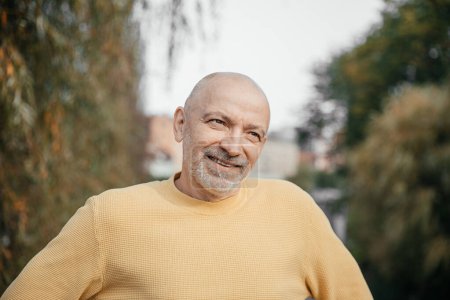 Elderly man in a yellow sweater, smiling softly, enjoying the serene outdoor environment in a lush urban park