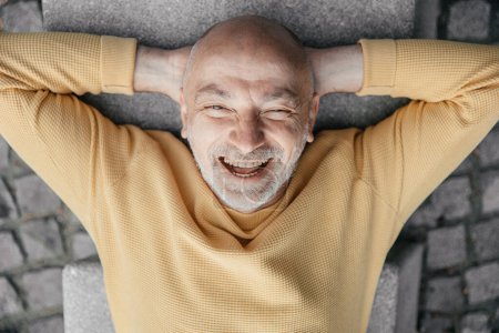 Homme âgé ludique allongé sur le dos sur le banc urbain faisant des visages drôles