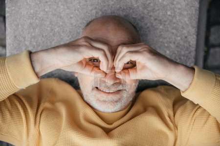 Joyful Senior Man Making Heart Shape with Hands While Lying on Urban Bench