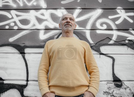 Senior man with a beard in a yellow sweater standing confidently with hands in pockets against a graffiti-decorated wall, looking upward