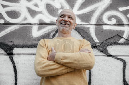 Cheerful bald senior man in a yellow sweater, arms crossed, standing against a vibrant graffiti wall, smiling broadly in an urban setting