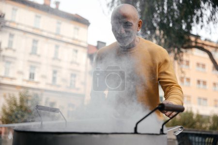 Senior man in a yellow sweater standing next to a large cooking pot on an urban street, smiling as steam rises around him
