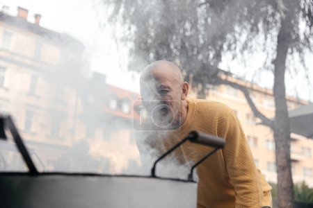 Senior man in a yellow sweater, focused and engaged as he checks the contents of a large steaming pot on an urban street