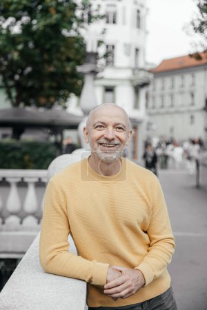 Hombre mayor sonriente en suéter amarillo apoyado en balaustrada urbana