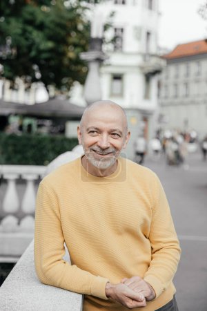 Smiling Senior Man in Yellow Sweater Leaning on Urban Balustrade