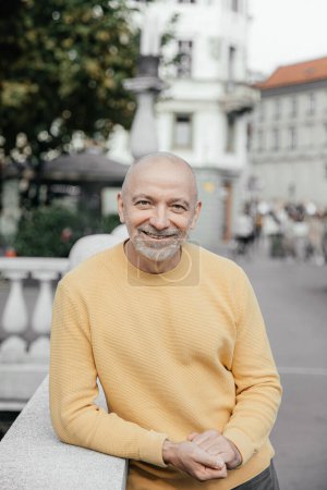 Fröhlicher, glatzköpfiger älterer Herr im gelben Pullover, gestützt auf eine steinerne Brüstung in urbaner Umgebung