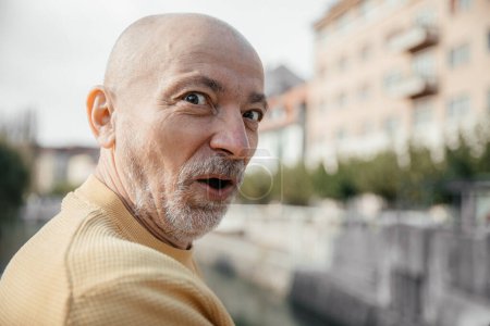 Ein älterer Mann im gelben Pullover zeigt einen überraschten Gesichtsausdruck, während er die Sehenswürdigkeiten entlang einer städtischen Uferpromenade genießt
