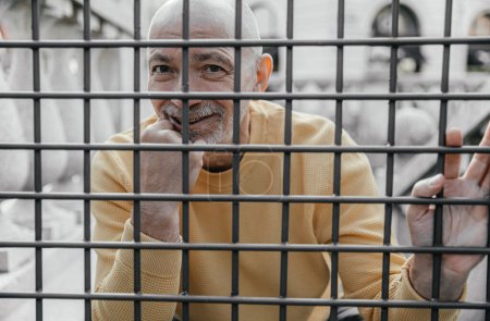Capturing a moment of contemplation, the elderly man appears thoughtful as he looks through metal bars, set in an urban environment