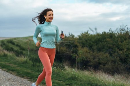 Photo for An attractive young woman is running in nature wearing fitness clothes and listening to music on her smartphone. - Royalty Free Image