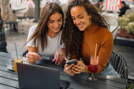 Téléchargez les photos : Deux filles sont assises dans le café, l'une regarde son téléphone intelligent pendant que l'autre vérifie les informations sur sa carte de crédit. - en image libre de droit