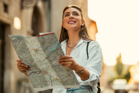Cheerful woman wanderer with trendy look searching direction on location map while traveling abroad in summer, happy female tourist searching road to hotel on atlas in a foreign city during vacation