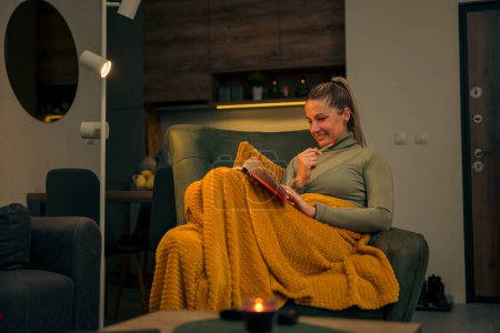 Téléchargez les photos : Belle jeune femme blonde assise dans un fauteuil et lisant un roman, elle sourit et apprécie la lumière des bougies - en image libre de droit