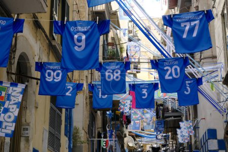 Foto de Camisas oficiales de SSC Napoli exhibidas en un histórico callejón de Nápoles, para celebrar el tercer campeonato de Nápoles, entre banderas y festones. - Imagen libre de derechos