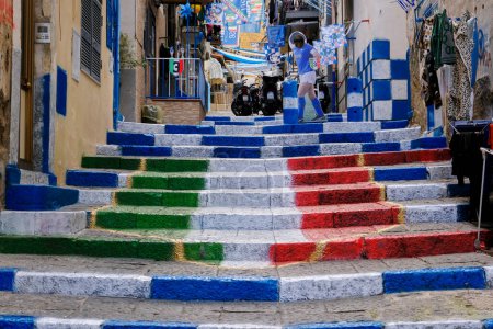 Foto de We are in Vico Rosario in Portamedina in the Montecalvario neighborhood in Naples. The steps of the alley have been painted with a tricolor shield and white and blue on the sides. In the background a life-size silhouette of Diego Armando Maradona. - Imagen libre de derechos