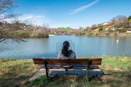 Foto de Mujer sentada relajada en un banco de jardín frente al lago Alain Cami en Saint Pee sur Nivelle, País Vasco - Imagen libre de derechos